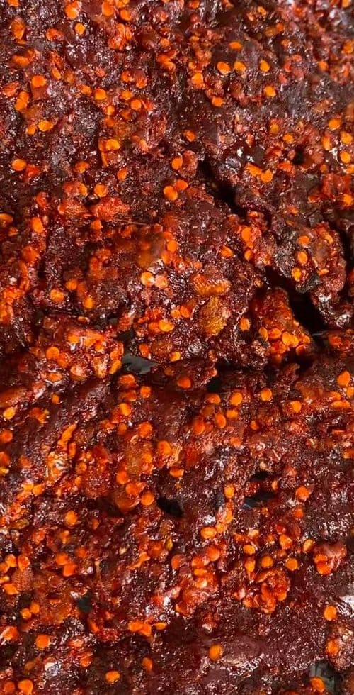 A gloved hand holding a torn, chili-flecked slab of kilishi